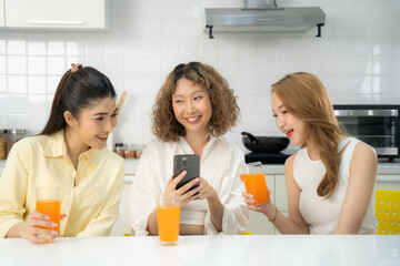 Three Asian women enjoying a light-hearted conversation with orange juice in a modern white kitchen, one using a smartphone.