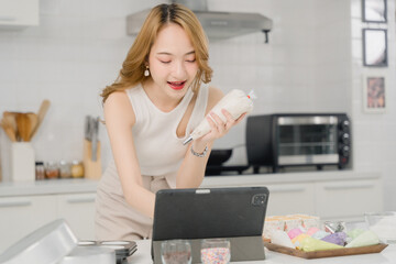 Smiling woman decorates cake with icing while following an online tutorial on a tablet in a modern kitchen.
