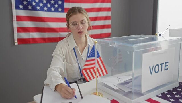 A young caucasian woman with glasses serving as a poll worker at a usa electoral voting station indoors, with an american flag backdrop.
