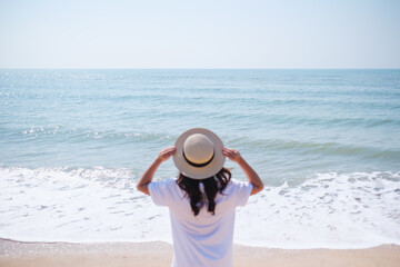Rear view image of a young woman with hat standing on the beach with blue sky background