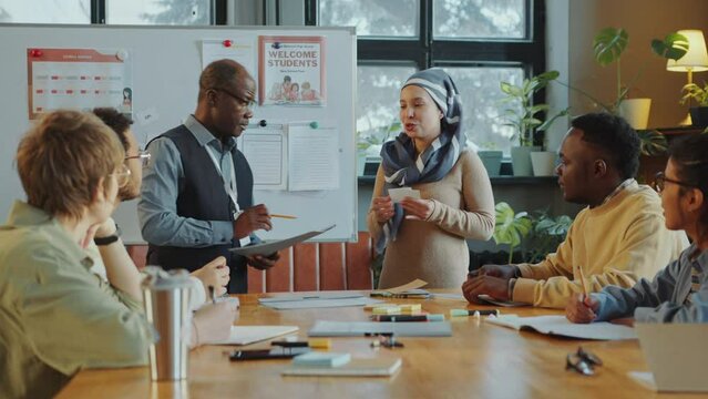Young Muslim woman in hijab standing by whiteboard with Black teacher, showing word cards and explaining them to group of immigrant students on lesson in language school