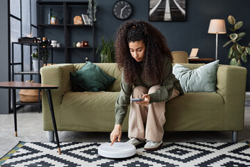 Long shot of young biracial woman sitting on sofa in living room turning on robotic vacuum cleaner