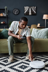 Fototapeta premium Vertical shot of young Black man sitting on sofa turning on modern robotic vacuum cleaner