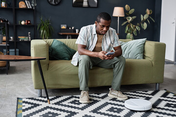 Long shot of African American man sitting on couch in modern living room adjusting robotic vacuum...