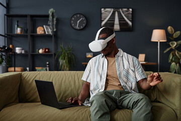 Young African American man sitting on sofa in living room setting VR headset using app on laptop,...