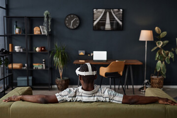 Rear view of unrecognizable Black man wearing VR headset sitting relaxed on couch in living room, copy space