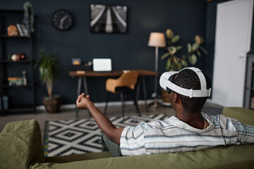 Rear view of unrecognizable young Black man sitting on couch in living room at home using VR...