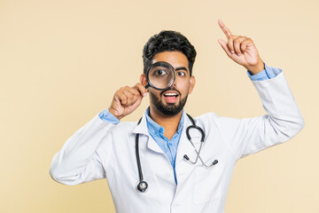 Investigator researcher scientist Indian young doctor cardiologist man holding magnifying glass near face looking , searching, analyzing. Arabian apothecary pharmacy guy isolated on beige background