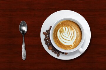 A cup of coffee with leaf design in the foam, surrounded by coffee beans and a spoon on the left.with beautiful dark brown wooden table background