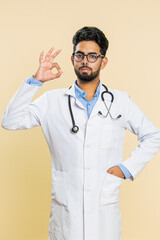 Okay. Happy Indian young doctor cardiologist man show ok gesture, like sign positive feedback, approve something good, celebrate victory. Arabian apothecary pharmacy guy on beige background. Vertical