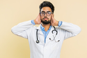 Don't want to hear and listen. Frustrated annoyed irritated Indian young doctor cardiologist man covering ears gesturing no, avoiding advice ignoring voices. Arabian apothecary guy on beige background