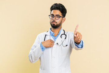 I am don't want it, not me. Confused Indian young doctor cardiologist man pointing fingers ask say who why me no thanks i do not need it rejection refusal stop sign. Apothecary guy on beige background