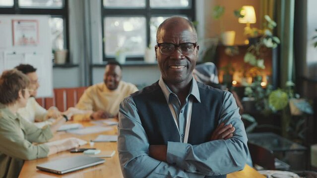 Senior African American teacher holding arms crossed and looking at camera with smile, group of immigrant students sitting behind in classroom. Video portrait