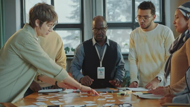 Group of multi-ethnic immigrant students and senior Black teacher standing around the table in classroom and discussing word cards during language class