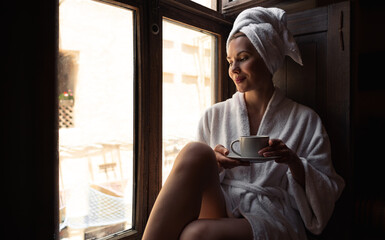 Woman in dark room sitting in robe by window. Attractive young girl in bathrobe and with towel on...
