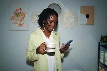 Smiling young Black businesswoman drinking cup of coffee and checking notifications on smartphone