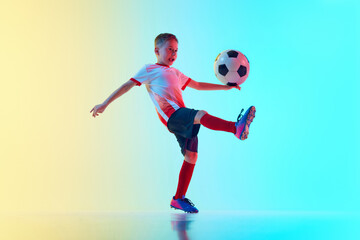 Dynamic shot of little boy in sports uniform plays football, training passing ball in neon light against gradient background. Concept of professional sport, championship, youth league, hobby. Ad