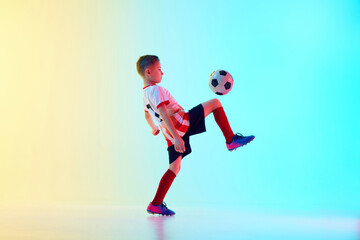 Dynamic photo of little athletic boy, soccer player kicking ball with knee in motion in neon light...