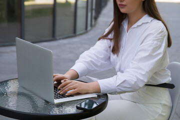 A woman in a white shirt and white pants sits at a table in a street cafe and works at a laptop.