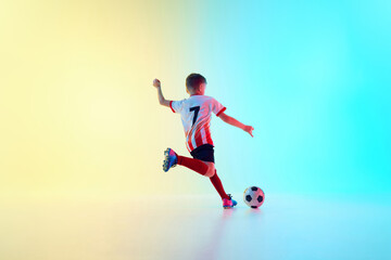 Young soccer player. Little athlete boy, dribbling ball to take perfect goal in neon light against gradient background. Concept of professional sport, championship, youth league, hobby. Ad