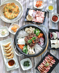 A korean food spread on a wooden table, featuring a central hot pot with noodles, vegetables, and slices of meat, surrounded by side dishes including dumplings, kimchi, and raw meats ready