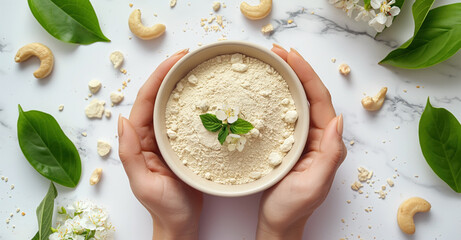 Hands holding a bowl of cashew powder surrounded by cashew nuts, green leaves, and white flowers. This image is set on a marble background, emphasizing the natural and healthy theme