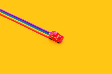 Blue toy car, rainbow ribbon flag trail road, yellow background. June gay parade, proud month, coming out day, human rights and tolerance concept. Minimal pop art composition. Flat lay, top view