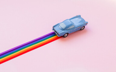 Blue toy car, rainbow ribbon flag trail road, pink background. June gay parade, proud month, coming out day, human rights and tolerance concept. Minimal pop art composition. Flat lay, top view