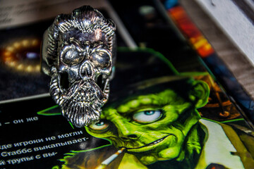 A ring with a skull, beard and mustache stands on a page with a painted zombie	
