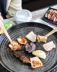 A person using chopsticks to grill various vegetables and meat on a round Korean BBQ grill at a restaurant table.