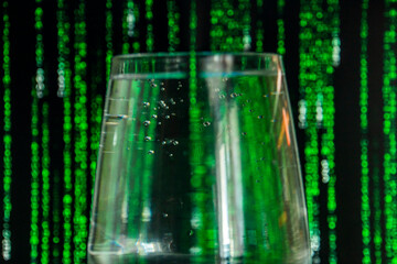 A transparent glass with bubbling water stands against the background of green falling numbers and letters close-up	
