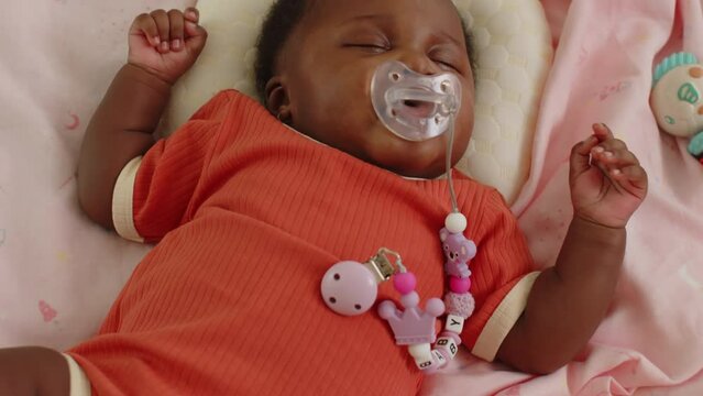 Handheld shot of adorable black baby girl in orange bodysuit lying in bed with eyes closed and sucking pacifier