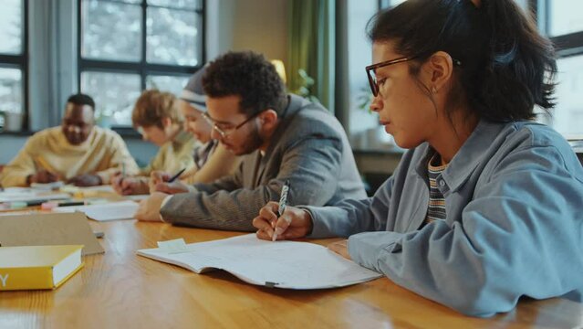Young Hispanic woman sitting at classroom desk with diverse group mates and writing in copybook when taking foreign language lesson in school for immigrants