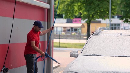 Caucasian handsome man washing his car using high pressure water and foam at self service wash station. Young guy cleaning auto outdoor at car cleaning point. Maintenance concept