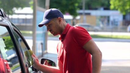 Portrait of Caucasian handsome man wiping the glass and the body of the car in car wash unit. Male...