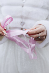 a bride in a white wedding dress and a white fur coat holds pink ribbons in her hands