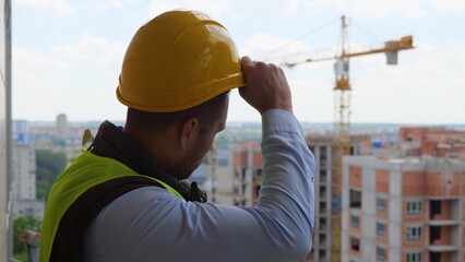 Close up portrait of Caucasian handsome professional male builder in uniform standing at construction site and taking off helmet while looking away. Building industry concept