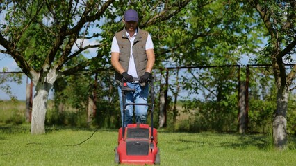 Male worker working in private garden mowing lawn with roller machine outdoors. Man walking in yard...
