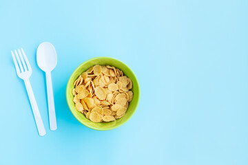 Corn flakes in green bowl with spoon and fork on blue background, Breakfast cereal