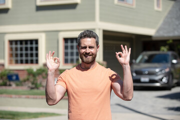 Naklejka premium Man with ok outdoor near house and car. Happy business man posing outside. Businessman. Real estate agent show ok sign. Business man in t-shirt. Car or house owner. American owner, casual businessman.