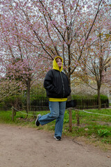 A woman in a yellow hood enjoys a playful moment, balancing on one foot while walking under the stunning pink blossoms of sakura trees