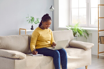 Happy woman enjoying online education in comfort of her own home. Young African American student girl sitting on sofa, using laptop computer, studying new useful information, and taking notes