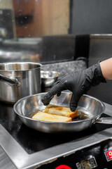 A close-up of a hand wearing a black glove, holding a piece of food being fried in a pan, with a pot nearby on the stove