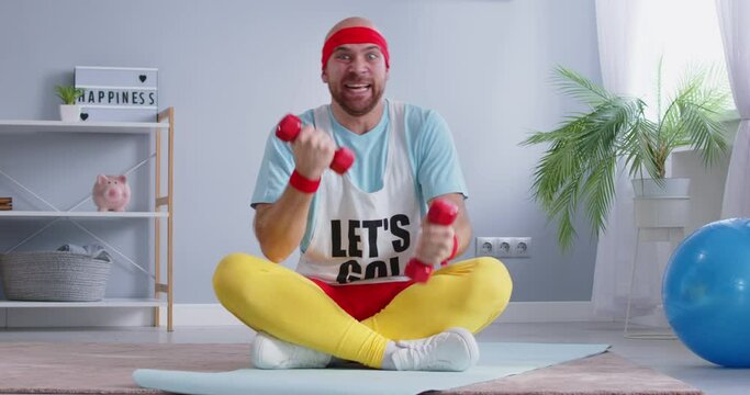 Humorous nerdy man in 80s sportswear is exercising with dumbbells at home, quickly doing bicep sport exercise while sitting on the floor. He dedicated to workout, blending retro style with fitness.