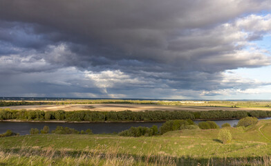 Naklejka premium moment in a rural landscape, featuring a wide river meandering through fields and pastures. The horizon is dominated by a dramatic sky filled with storm clouds