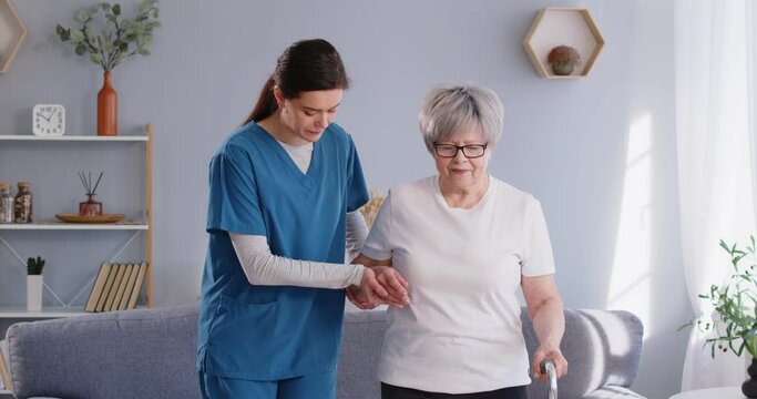 In home setting, female doctor or nurse support senior patient in rehabilitation, aiding them in walking with cane at home living room. Care and supportive presence enhance patient ability to walk.