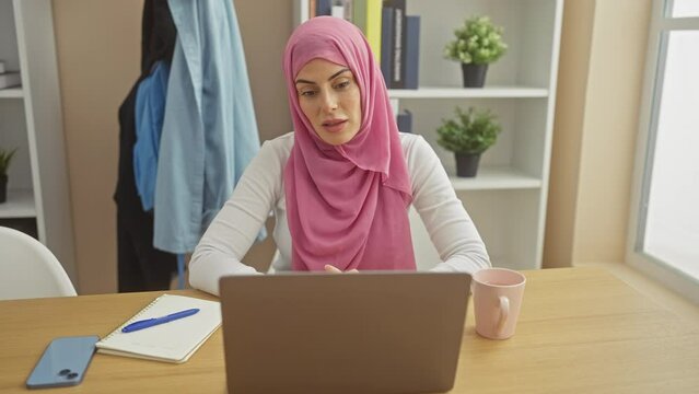 Stressed young hispanic woman, adorning a hijab, grappling with frustration and anxiety as she works on a computer indoors, her living room echoing her shock, anger and worry.