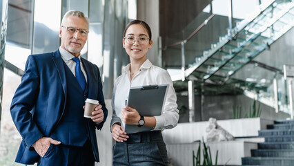 Portrait of mature smiling business man and business woman standing posing in office. Two diverse colleagues, group team of confident professional business people.