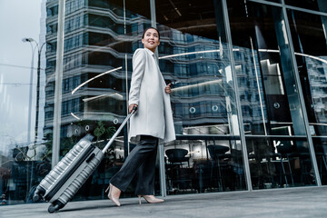 Beautiful young businesswoman with suitcase walking outside airport. Woman travel to hotel or office. Portrait of attractive professional walking in city street with bag