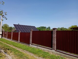 Solar panels are mounted behind a tall red sheet metal fence to generate solar energy into electrical energy. The topic of using solar energy for human needs. Electricity generation from renewable nat